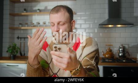 Mann, der in der Küche sitzt, reagiert auf das, was sieht, surft im Internet auf dem Handy, liest E-Mails, liest Mediennachrichten, scrollt in sozialen Medien, benutzt Mobil Stockfoto