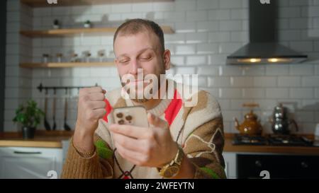 Der Mann sitzt in der Küche und reagiert auf das, was er sieht: Er surft im Internet auf dem Mobiltelefon, liest E-Mails, liest Mediennachrichten, scrollt in sozialen Medien und verwendet mobile ap Stockfoto