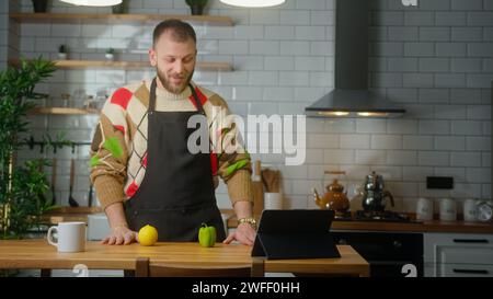 Vlogger Chef auf einer Schürze spricht in Tablet-Computer, nimmt Video-Kochkurs über vegetarische Mahlzeiten oder Salate in der modernen Küche zu Hause auf. Online Stockfoto