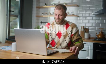 Mann, der zu Hause in der Küche arbeitet, mit seinem Laptop arbeitet, Notizen macht, recherchiert. Viel beschäftigter Freiberufler, der an modernen technischen Notebooks arbeitet Stockfoto