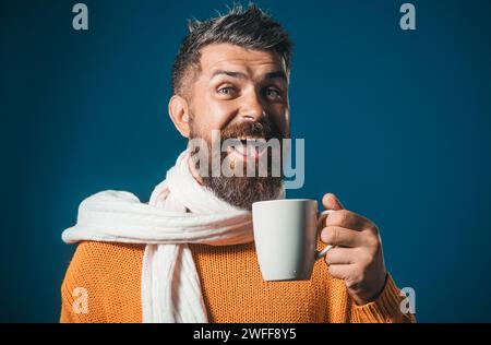 Glücklicher bärtiger Mann in orangefarbenem Pullover und weißem Schal genießt eine Tasse Kaffee oder Tee im Café. Nahaufnahme Porträt eines gutaussehenden Mannes in modischer Kleidung Stockfoto