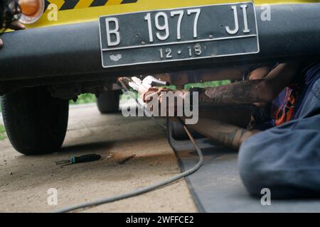 Ein junger Mann, der ein Lenkproblem an einem alten Auto repariert, unter dem Auto schweißt. Stockfoto