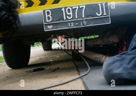 Ein junger Mann, der ein Lenkproblem an einem alten Auto repariert, unter dem Auto schweißt. Stockfoto