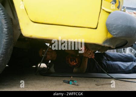 Ein junger Mann, der ein Lenkproblem an einem alten Auto repariert, unter dem Auto schweißt. Stockfoto