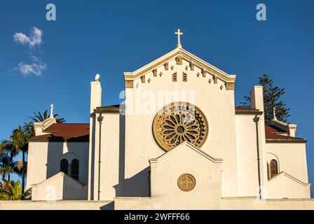 Santa Barbara, CA, USA - 19. Januar 2024: Beige Südfassade der Kirche Our Lady of Sorrows unter Sonnenlicht vor blauem Himmel. Grünes Laub hinten. Stockfoto