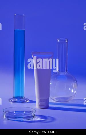 Weißes Röhrchen mit leerem Etikett, das mit einem Röhrchen, einer Petrischale und einem florenzkolben mit flachem Boden angeordnet ist. Kosmetisches Produkt-Mockup mit Laborkonzept Stockfoto