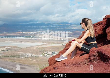 Eine junge Frau findet Trost und Besinnung, während sie auf einem markanten roten Felsen sitzt und auf den weiten Ozean blickt. Stockfoto