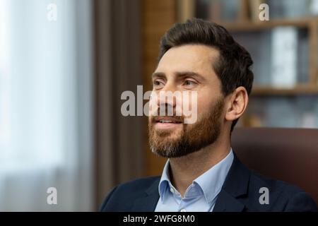 Selbstbewusster kaukasischer Mann mit trendiger Frisur, die nach vorne blickt und auf unscharfem Hintergrund inspiriert ist. Zufriedener männlicher Vorgesetzter, der sich den Quartalsbericht mit beeindruckenden Ergebnissen des eingestellten Mitarbeiters im Kabinett anhört. Stockfoto