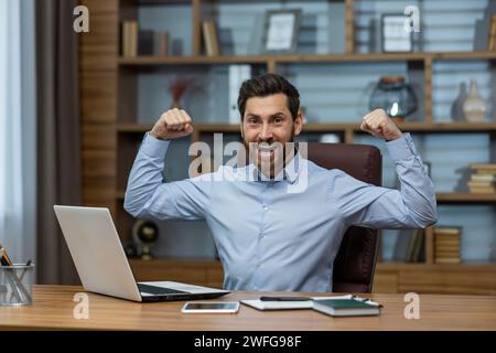 Ekstatischer bärtiger Mann in blauem Hemd, der die Hände mit geballten Fäusten in Siegerposition hebt, während er am Computer arbeitet. Freudiger Unternehmer, der sich freut, während er schreiend und erfolgreich feiert. Stockfoto