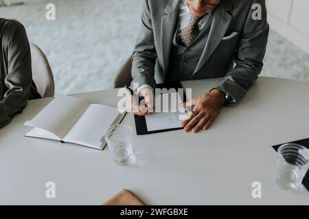 Mittelteil eines Reifen Geschäftsmannes, der in Papier schreibt, während er während des Meetings am Konferenztisch sitzt Stockfoto