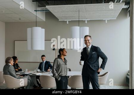 Lächelnde, reife Geschäftsfrau, die neben einem jungen Geschäftsmann mit einer Tasse Kaffee im Sitzungsraum steht Stockfoto