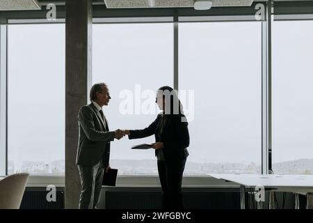 Geschäftsmann und Geschäftsfrau schütteln die Hände, während sie am Fenster im Büro stehen Stockfoto