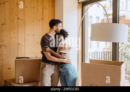 Glückliches Paar, das sich umschließt, während es in der Nähe des Fensters in einem neuen Zuhause steht Stockfoto