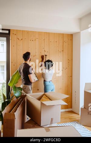 Frau hämmert Nagel auf Holzwand, während sie mit Freund in einem neuen Zuhause steht Stockfoto