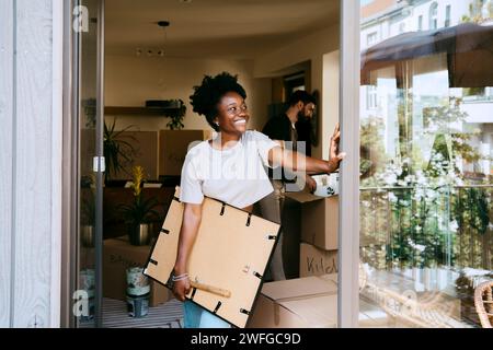 Lächelnde Frau, die einen Rahmen hält, während sie sich in einem neuen Zuhause an die Wand lehnt Stockfoto