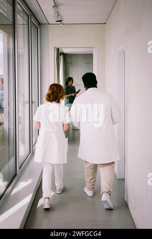 Rückansicht der Oberärztin in voller Länge, die mit einem männlichen Kollegen im Spitalflur läuft Stockfoto