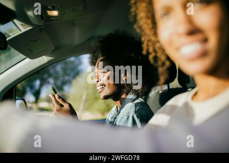 Lächelnde junge Frau mit Smartphone, die bei einem Freund im Auto sitzt Stockfoto
