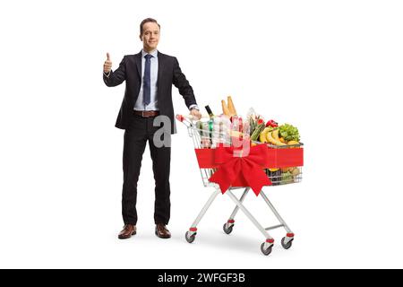 Porträt eines Geschäftsmannes in voller Länge, der Daumen nach oben zeigt mit Essen in einem Einkaufswagen, Food Charity Konzept isoliert auf weißem Hintergrund Stockfoto