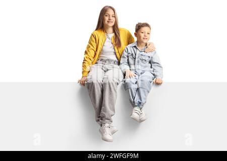 Teenager-Schwester mit ihrem jüngeren Bruder, der auf einem leeren Panel sitzt und in die Kamera lächelt, isoliert auf weißem Hintergrund Stockfoto