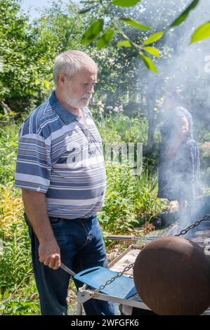 Ein grauhaariger Mann grillt Fleisch auf einem Grill beim Brazier. Fleisch wird auf einem hausgemachten Grill in der Natur zubereitet. Stockfoto