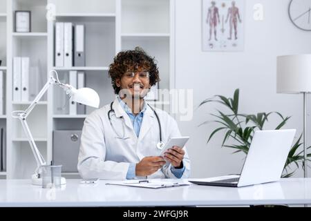Fröhlicher junger männlicher Arzt mit Stethoskop, der an seinem Schreibtisch in einem medizinischen Büro sitzt, an einem Laptop arbeitet und ein Tablet hält. Stockfoto