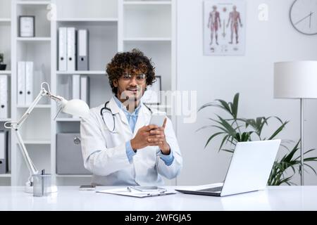 Professioneller junger hinduistischer männlicher Arzt mit lockigen Haaren, im medizinischen Mantel, mit einem Smartphone in einer gut ausgestatteten Klinik. Stockfoto