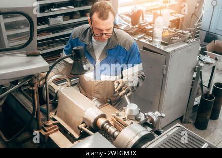 Porträt des professionellen turners bei der Arbeit an der Drehmaschine in der Werkstatt. Der 50-55-jährige turner in Overall und Brille dreht das Teil in der Werkstatt an der Maschine. Fotografie authentischer Arbeitsprozess. Stockfoto