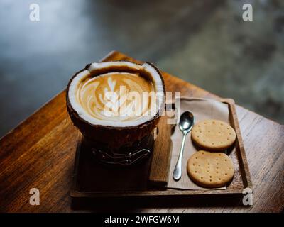 Heißer Latte-Kaffee in Kokosnuss. Tablett mit einer Tasse leckeren Kokoskaffee. Tasse leckeren Kokosnusskaffee und Löffel mit Crackern auf Holztisch. Warmes Café lat Stockfoto