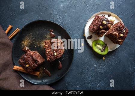 Blick von oben auf vegane Schokoladen-Brownie auf Stoffserviette mit geschnittenen Kiwis auf dunklem, stimmungsvoll gemalten Hintergrund Stockfoto