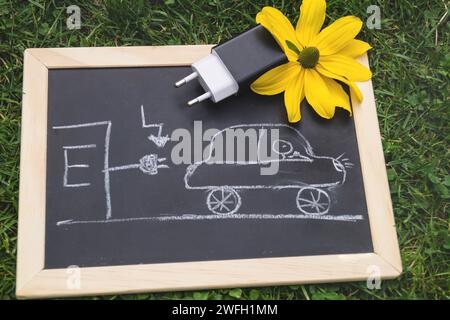 Schwarze Tafel mit einem gezogenen Elektroauto an einer Ladestation, Stecker und gelbe Blüte, Symbolbild Stockfoto