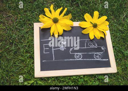 Schwarze Tafel mit einem gezogenen Elektroauto an einer Ladestation und gelben Blüten, Symbolbild Stockfoto