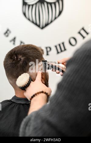 Barber rasiert die Kontur der ovalen Linie mit einem Haarschneider am Kopf des Kunden. Ein Mann mit Bart bekommt einen Haarschnitt in einem Friseurstuhl. Stockfoto