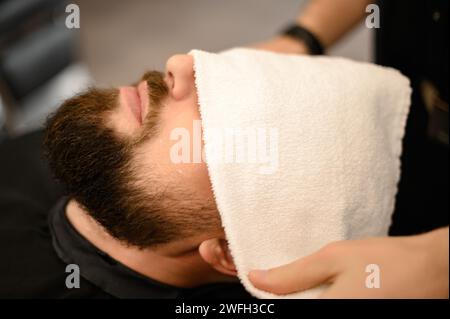 Der Friseur bedeckt das Gesicht des Kunden mit einem feuchten Handtuch, um den Bart abzudampfen. Stockfoto