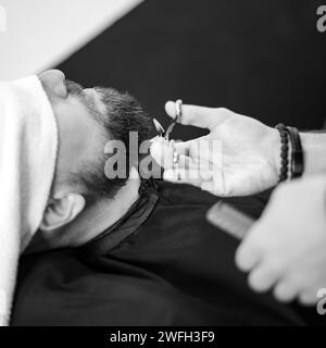 Ein Friseur beschneidet den Bart eines Kaukasiers, dessen Gesicht mit einem Handtuch bedeckt ist, mit einer Schere Stockfoto