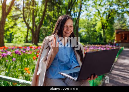 Außenporträt einer glücklichen Geschäftsfrau, die im Frühlingspark auf einem Laptop arbeitet. Frau ruft durch Videokonferenzen an und winkt Cowokern im öffentlichen Garten zu Stockfoto