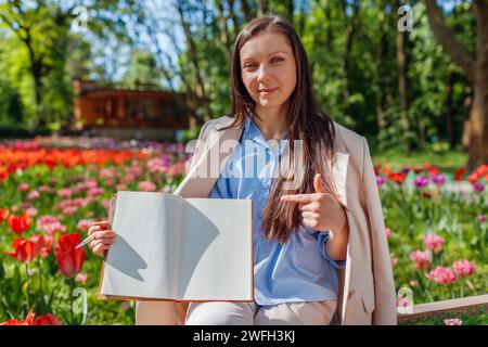 Porträt einer jungen kaukasischen Geschäftsfrau, die auf geöffnetes Notizbuch mit leeren Seiten im Frühlingspark zeigt. Kopierbereich. Modell Stockfoto
