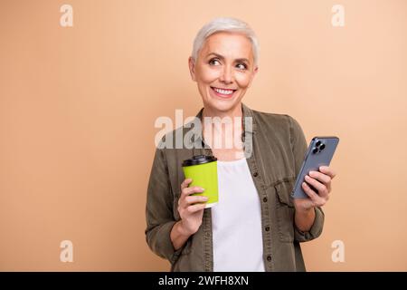 Foto von glücklichem grauem Haarmodell Geschäftsfrau mit Kaffeetasse Genießen Sie das Trinken von Cappuccino, bevor Sie isoliert auf beigefarbenem Hintergrund arbeiten Stockfoto