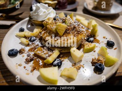 Ein Stapel Pfannkuchen mit frischen Apfelscheiben, Heidelbeeren, knusprigem Granola, einem Staubbeutel Puderzucker und einer Seite Schlagsahne. Stockfoto