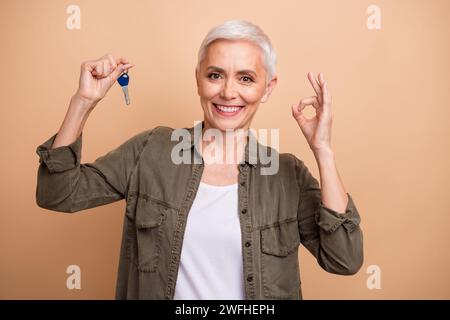 Foto-Porträt der attraktiven pensionierten Frau halten Schlüsselshow okey Symbol gekleidet stilvolle Khaki Kleidung isoliert auf beigefarbenem Hintergrund Stockfoto