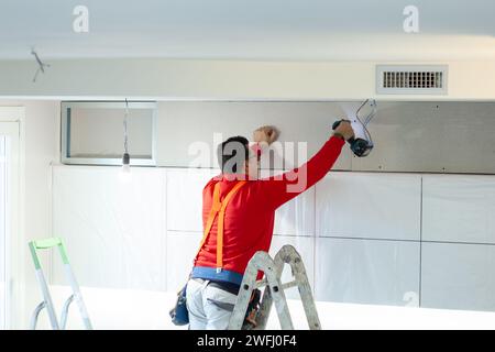 Ein Arbeiter aus Gipskarton installiert eine Gipskartonwand an den Küchenschränken, um das Absaugrohr der Haube abzudecken. Stockfoto