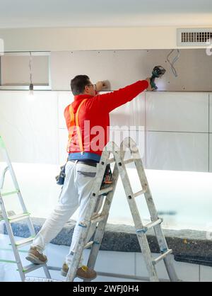 Ein Arbeiter aus Gipskarton installiert eine Gipskartonwand an den Küchenschränken, um das Absaugrohr der Haube abzudecken. Stockfoto