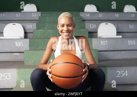 Eine afroamerikanische Basketballspielerin, die auf den Stadionständern sitzt, lächelnde Frau, die einen Basketball hält, verkörpert das Konzept der Leidenschaft für die s Stockfoto