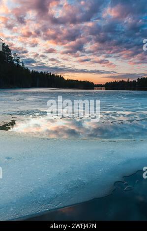 Eine ruhige Szene fängt die Schönheit eines teilweise gefrorenen Sees in der Abenddämmerung ein, mit subtilen Farbtönen des Sonnenuntergangs, die auf dem Eis und dem Wasser im s reflektieren Stockfoto