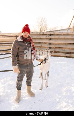 Vertikale Aufnahme eines hübschen, glücklichen jungen Mannes in warmen Kleidern, der niedliche junge Rentiere auf einer verschneiten Hirschfarm am sonnigen Wintertag umarmt und füttert Stockfoto