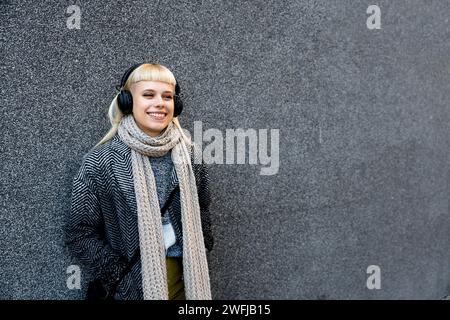 Junge, schöne Hippie-Hipster-Frau in moderner Kleidung, die schnurloses Headset trägt und Musik hört, während sie sich an die Wand lehnt. Blonde Frau mit coolem Attitu Stockfoto