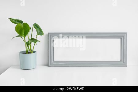 Grauer Mock Up Bilderrahmen und Monstera deliciosa oder Schweizer Käsepflanze in einem Blumentopf auf weißem Tischhintergrund Stockfoto