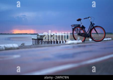 Holland, Zeeland - 09. Juni 2022: Hafengebäude von Rotterdam am Horizont bei Sonnenaufgang. Stelzenhaus am Ufer der Nordsee. Damen Fahrrad i Stockfoto