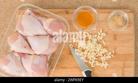 Plastikbehälter mit rohen Bio-Hühnerstäbchen, gehacktem Knoblauch auf einem Holzschneidebrett, Kurkuma und Gewürzen in kleinen Schüsseln, Nahaufnahme auf A Stockfoto