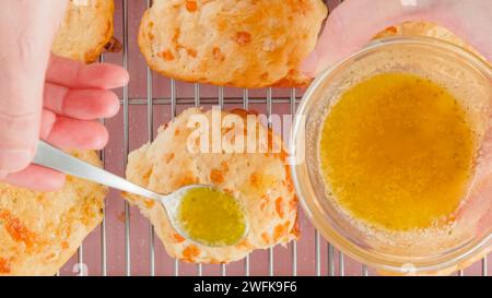 Käsegebäck Rezept, Backprozess. Frau, die frisch gebackene Kekse mit geschmolzener Butter und Knoblauchwürz garniert, flach gelegt Stockfoto