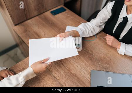 Draufsicht einer Geschäftsmanagerin mittleren Alters, die Papiere an eine nicht erkennbare Kollegin weitergibt, die am Tisch im Büro sitzt. Stockfoto